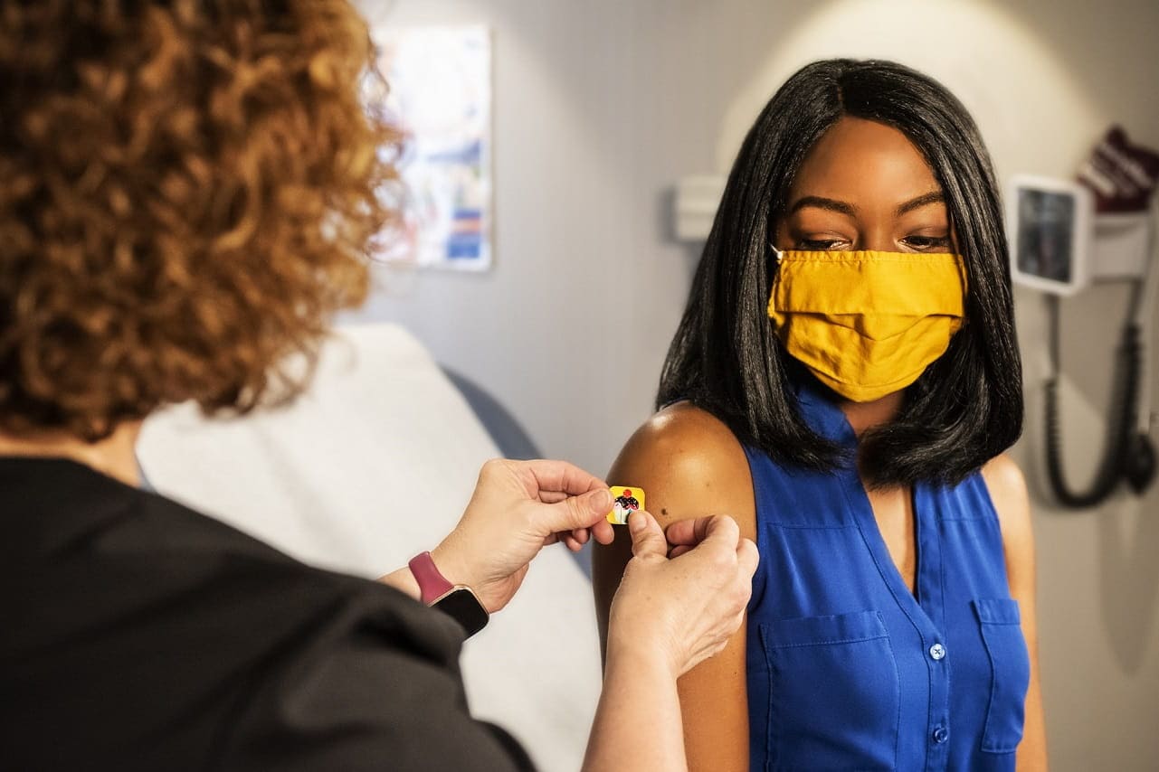 Woman in blue shirt with mask on getting a band-aid on upper arm after a vaccination.