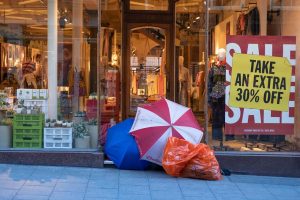 A homeless person camped in front of a clothing store with Sale signs in the window.